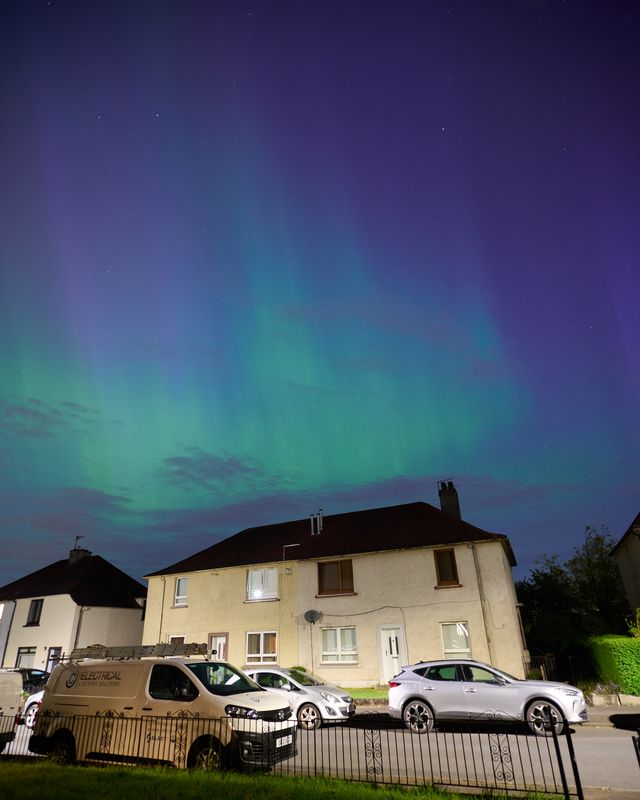 A street with parked cars and a wall of cyan aurora above the buildings.